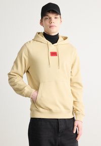 Sudadera con capucha beige y bolsillo tipo canguro, que presenta un parche de logo rectangular rojo. El modelo lleva una gorra negra y un cuello alto debajo.