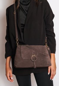 Donkerbruine suedine crossbody tas met een voorflap, ronde hardwareaccent en verstelbare schouderband. Het oppervlak is zacht met een gestructureerde afwerking.