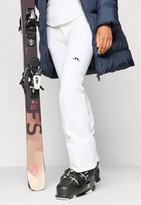 Pantaloni de schi albi cu o textură netedă, bocanci de schi negri cu accesorii argintii și o jachetă puffer albastră, ținând o placă de snowboard, parțial vizibilă.