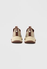 Bežā un brūnā krāsā slip-on stila kedu pāris ar spīdīgu akmentiņu rotājumu, skatīts no aizmugures uz vienkārša pelēka fona.