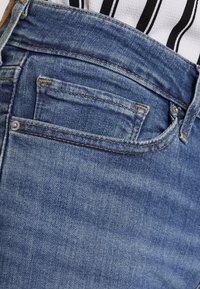 Jean en denim bleu avec une coupe classique, un design à cinq poches, et une texture subtilement délavée avec des surpiqûres jaunes contrastantes.