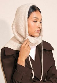 Be�żowa dziana czapka z miękkiego materiału, z prążkowaną fakturą, w połączeniu z brązowym płaszczem trench, z białym golfem pod spodem.