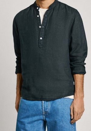 Hombre con una camisa henley de manga larga verde oscuro sobre una camiseta blanca y pantalones vaqueros azul claro, de pie frente a un fondo claro.