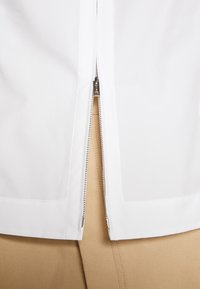 Fermeture éclair argentée partiellement ouverte sur une veste blanche, révélant un pantalon beige texturé en dessous.