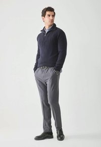 Jersey de punto azul marino con cuello de cremallera, pantalones grises a medida y zapatos negros; presenta un patrón texturizado en la parte frontal del jersey.