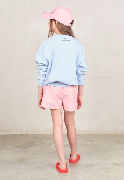 Felpa azzurra, pantaloni corti rosa e ciabatte rosse. Presenta un logo sul retro della felpa e dettagli con bottoni sui pantaloni corti.