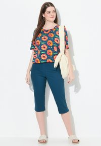 Haut à imprimé floral avec de grandes fleurs orange et roses sur un fond marine, associé à un pantalon capri teal et des sandales légères. Comprend un sac beige.