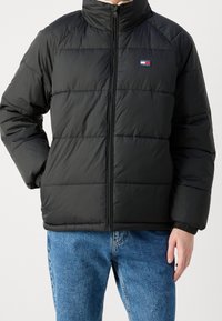 Mężczyzna w czarnej, pikowanej kurtce zapinanej na zamek, z logo Tommy Jeans, w połączeniu z niebieskimi dżinsami.