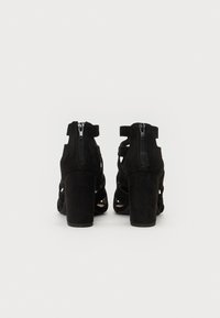 Zwarte suede hoge hakken met een open ontwerp, voorzien van achteraan ritsen en een chunky hak, die uitsnijdingen en een gladde textuur benadrukken.