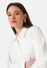 White collared shirt with floral embroidery and cut-out patterns, paired with silver abstract earrings. Smooth texture, button closure visible.