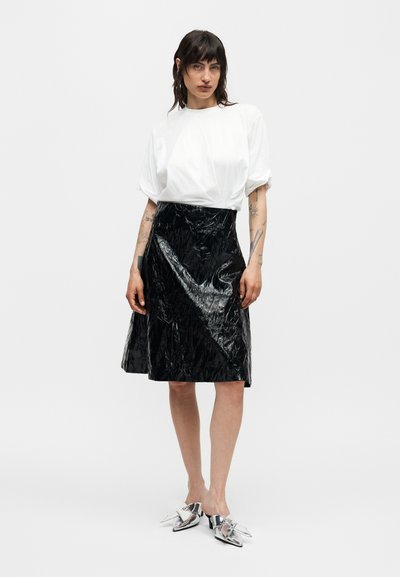 Γυναίκα που φοράει άσπρο άνετο κοντομάνικο τοπ, γυαλιστερή μαύρη φούστα μέχρι το γόνατο και μεταλλικά μυτερά παπούτσια χωρίς κορδόνια, στέκεται μπροστά σε απλό φόντο.