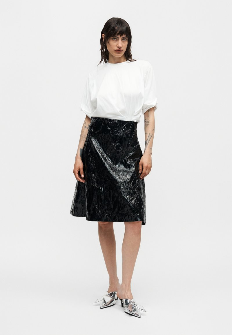 Kvinde iført en løs hvid kortærmet top, skinnende sort knælængde nederdel og metalliske spidse slip-on sko, står foran en ensfarvet baggrund.