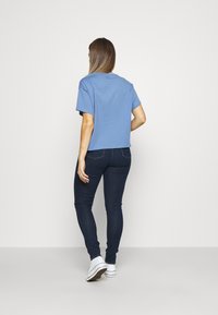 Mujer con una blusa de manga corta azul claro, jeans oscuros ajustados y zapatillas blancas, de pie con la espalda hacia la cámara.