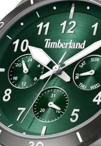 Okrogla ura Timberland z temno zelenim metalnim številčnikom, srebrnimi kazalci in oznakami, tremi podštevilčnicami ter teksturirano površino.