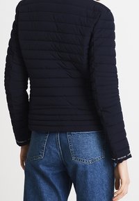 Navy quilted jacket with a fitted design and long sleeves, featuring horizontal stitching and logo accents on the cuffs.