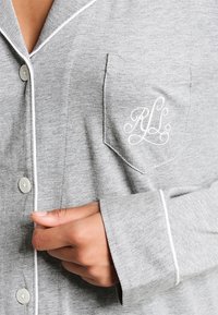 Camisa de pijama cinza feita de um tecido suave, apresenta um acabamento branco ao longo do colarinho e das mangas, com abertura frontal por botões e um bolso com iniciais bordadas.