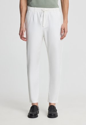 Uomo che indossa pantaloni bianchi con coulisse e scarpe slip-on nere, in piedi davanti a uno sfondo chiaro e uniforme.