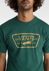 Camiseta de algodón verde con un gran gráfico dorado que presenta la palabra "VANS" y un diseño de patineta, también mostrando "desde 1966". Cuello redondo.