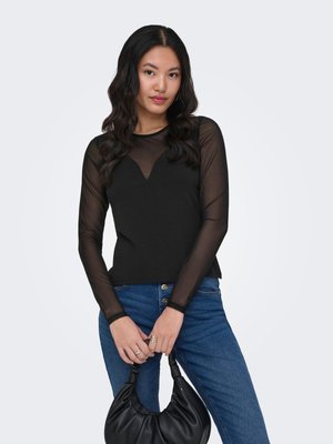 Femme aux longs cheveux noirs portant un haut noir transparent à manches longues et un jean bleu, tenant un sac à main noir sur un fond uni.