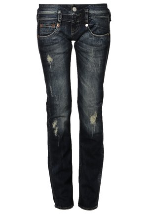 Persona che indossa una maglia a maniche lunghe a righe bianche e nere, jeans scuri consumati risvoltati alle caviglie, sneaker blu navy e porta uno zaino in pelle marrone.