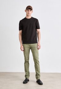 Rjava majica s kratkimi rokavi, svetlo zelene hlače slim fit in črne natikače. Model nosi usklajen rjav klobuk. Preprost, minimalističen dizajn.
