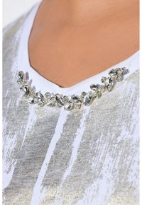 Maglietta bianca con un motivo a strisce argentate glitterate. Presenta un colletto decorativo con strass di varie forme. Tessuto morbido.