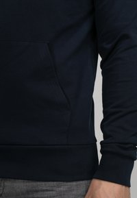 Marineblaue Sweatshirt mit langen Ärmeln, weicher Textur, Kängurutasche sowie gerippten Bündchen und Saum. Nahaufnahme des Stoffes und der Designdetails.