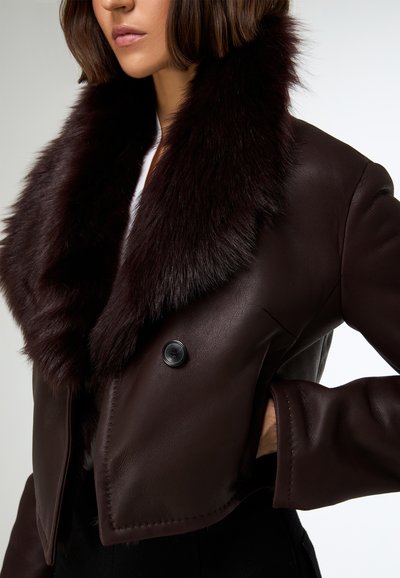 Giacca in pelle marrone scuro tagliata, con un ampio colletto in pelliccia sintetica, dotata di un singolo bottone nero e cuciture dettagliate lungo l'orlo.