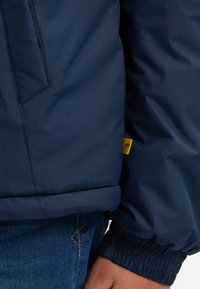 Marineblaue Steppjacke aus strukturiertem Stoff mit elastischen Bündchen und einem sichtbaren Logoetikett in Gelb. Gesteppte Details am Saum und an der Tasche.