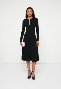 Čierne šaty s dlhým rukávom a rozšírenou sukňou, s gombíkovým výstrihom a hladkou textúrou. Nosí sa s čiernymi lodičkami a bielou kabelkou.