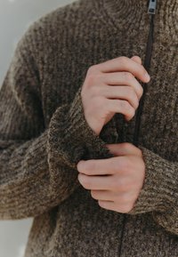 Bruine cardigan met ritssluiting en een geribbelde textuur, met nauwsluitende mouwen en een hoge kraag. Handen passen de mouw aan.