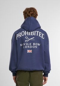 Navy blauwe hoodie met een trekkoordcapuchon. Witte tekst "PROHIBITED" en "SAVILE ROW LONDON" op de achterkant, met scharen en een UK-vlag.