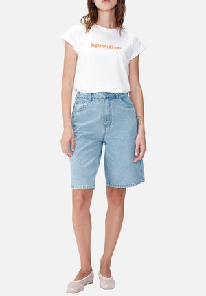 Femme portant un t-shirt blanc « aperitivo », un short en denim bleu clair longueur genou et des ballerines beiges, debout sur un fond blanc.