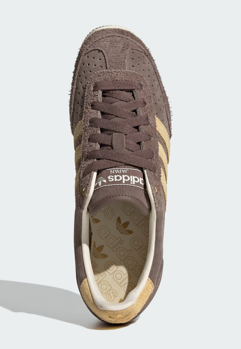 Brun mockasneaker med beige tre-randigt mönster. Har rundad tå, snörning och präglad logotyp på tungan. Texturerad innersula.