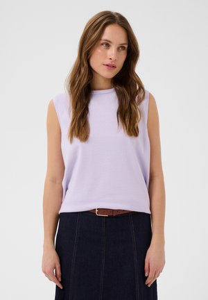 Femeie cu păr lung și castaniu care poartă un top mov deschis fără mâneci, băgat într-ună fustă închisă la culoare din denim cu o centură maro, stând pe un fundal simplu.