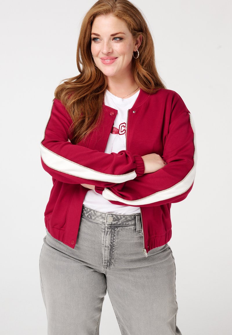 Veste bomber bordeaux avec des manches à rayures blanches. Matière douce, poignets ajustés et fermeture éclair ; superposée sur un t-shirt graphique blanc.