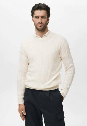 Suéter de punto trenzado en color crema claro con cuello redondo, patrón texturizado y dobladillo acanalado, usado sobre una camisa blanca con cuello y pantalones negros.