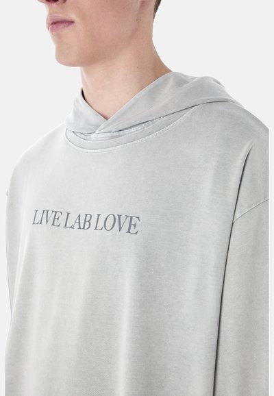 Homme portant un sweat à capuche gris clair avec l'inscription "LIVE LAB LOVE" en lettres majuscules sur la poitrine, vu des épaules au menton.