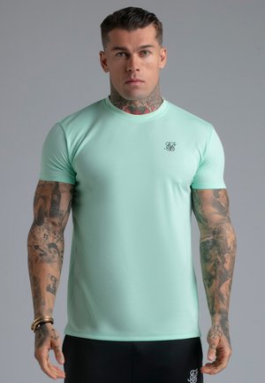 Man with tattoos wearing mint green fitted t-shirt and black shorts against plain gray background, facing forward with neutral expression.