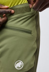 Χέρια που προσαρμόζουν τη μέση ενός πράσινου παντελονιού με κλείσιμο με αυτοκόλλητο βέλκρο, με τσέπη με φερμουάρ και λευκό λογότυπο μαμούθ κοντά στο τελείωμα.