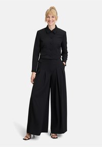 Zwarte cropped jas met een kraag en knoopsluiting, gecombineerd met wijde broek met plooien. Glad materiaal, minimalistisch ontwerp.