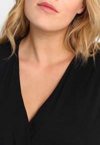 Czarna bluzka z dekoltem w serek, uszyta z miękkiego, gładkiego materiału, prezentująca luźny krój. Skóra widoczna w okolicy dekoltu podkreśla sposób opadania materiału.