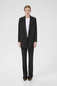 Svart dobbelspent blazer laget av glatt stoff, sammen med en hvit tettsittende topp og svarte bukser. Spisse olivengrønne hæler fullfører utseendet.