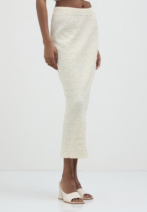 Donna che indossa una gonna midi a matita di crema con texture e mule aperte con tacco di crema, in piedi contro uno sfondo bianco semplice.