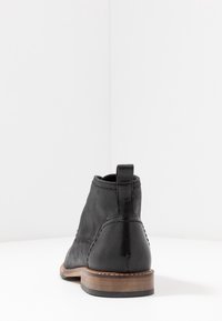 Botte noire en cuir jusqu'à la cheville avec une finition lisse, des coutures beiges et une semelle en bois marron. Elle présente une languette à l'arrière pour un enfilage facile.