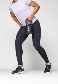 Una persona vestita con leggings neri e una maglietta viola tiene un kettlebell nero mentre esegue un affondo su uno sfondo grigio uniforme.