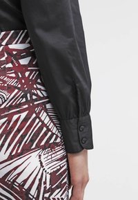 Un bras en chemise à manches longues noires avec un poignet boutonné reposant à côté d'une jupe ou d'un pantalon à motifs rouge, blanc et noir.