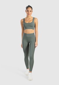 Olivově zelená sportovní podprsenka a legíny s vysokým pasem vyrobené z hladkého, pružného materiálu, doplněné o bílé sportovní boty.