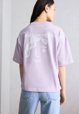 Γυναίκα που φοράει μια φαρδιά μπλούζα λεβάντας με το κείμενο "ΓΥΝΑΙΚΕΣ ΓΙΑ ΓΥΝΑΙΚΕΣ" και μια μινιμαλιστική γραφική εικόνα προσώπου στην πλάτη, συνδυασμένη με ανοιχτόχρωμο μπλε τζιν.