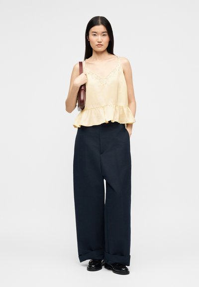 Femme aux longs cheveux noirs portant un caraco jaune à volants, un pantalon large bleu marine, des chaussures noires, et un sac à bandoulière marron.